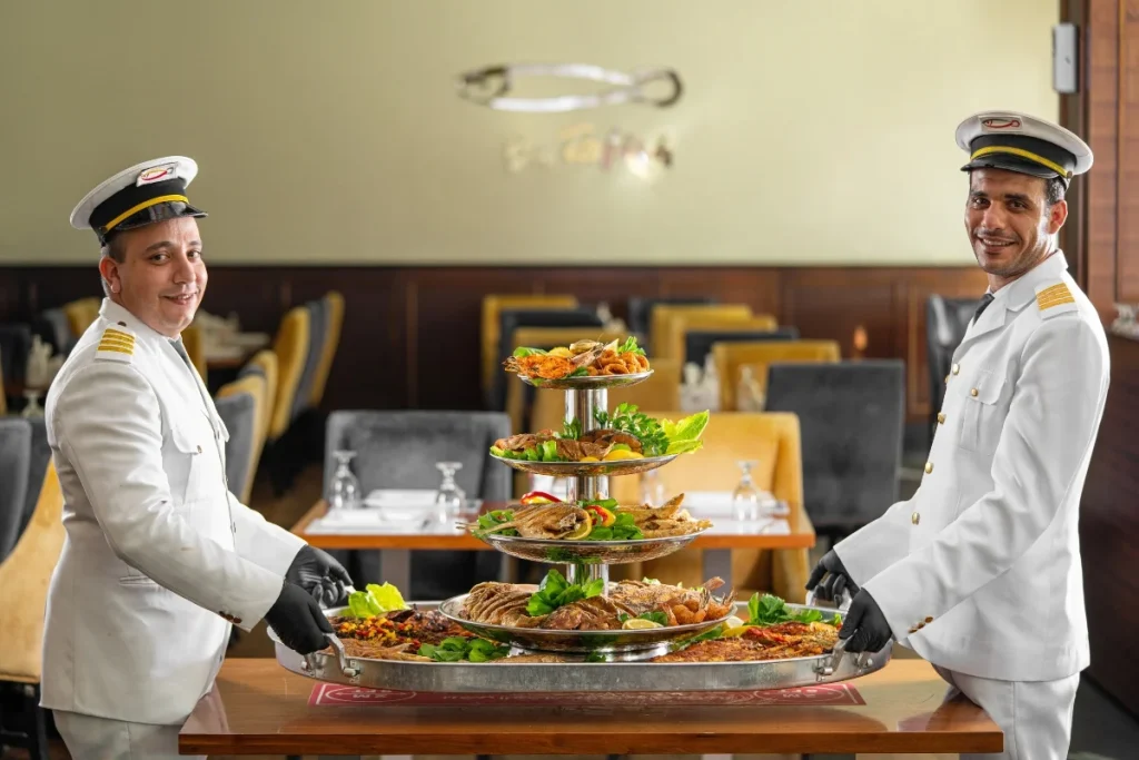 Two restaurant staff in classic uniforms present a large multi tier seafood platter inside an elegant dining space